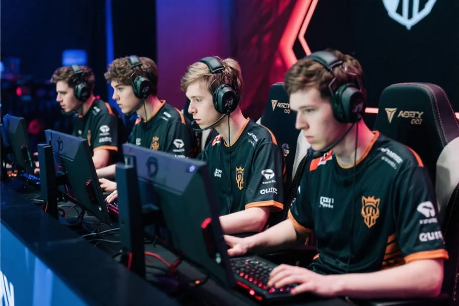 五位职业电子竞技选手正在聚精会神地操作电脑，身穿统一队服，背景为电竞赛事现场